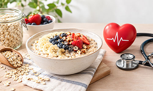 Semangkuk oatmeal dengan taburan beri segar, pisang, biji chia, dan kacang-kacangan di samping hati merah dan stetoskop, melambangkan manfaat oat bagi kesehatan jantung.