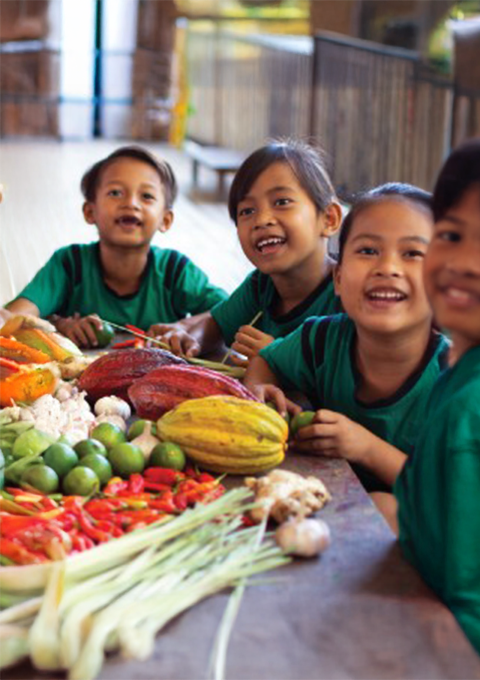 Anak-anak tersenyum di depan meja berisi kakao, buah, sayuran, dan rempah lokal.